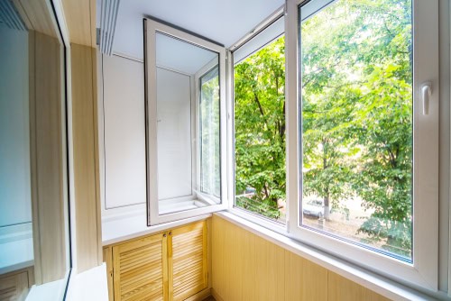 A bright enclosed balcony with large windows and lush green trees visible outside. The windows, installed by a top window installation expert, are open, allowing fresh air to enter. The balcony has a light wood-paneled lower wall and cabinet, creating a cozy atmosphere.