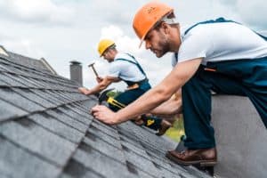 Two construction workers in hard hats are busy with a roof repair in Suffolk County. One worker in the background is using a hammer, while the worker in the foreground is placing shingles. Both are wearing white shirts and overalls as they work under a cloudy sky.