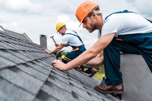 Two construction workers in hard hats are busy with a roof repair in Suffolk County. One worker in the background is using a hammer, while the worker in the foreground is placing shingles. Both are wearing white shirts and overalls as they work under a cloudy sky.