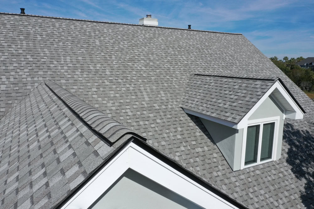 A close-up view of a house with a newly installed shingle roof by a general contractor in Suffolk County. The roof, featuring a gable design with a small dormer, is complemented by uniform gray shingles. A clear sky with few wisps of clouds provides the perfect backdrop.