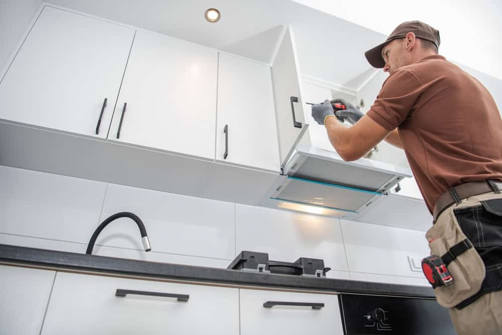A general contractor wearing a cap and gloves is installing or fixing a white kitchen cabinet with a drill. The modern kitchen features white cabinets, a black countertop, a black faucet, and a stovetop. He is standing on a step ladder and appears focused on his task.