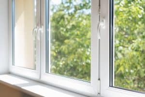 An image of a white-framed window with two visible panels. The window is open, allowing a view of lush green foliage outside. Sunlight is streaming in, creating a bright and airy atmosphere inside. The window handles are white and positioned vertically—courtesy of expert window installation.