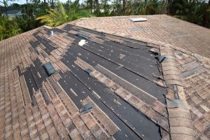 A rooftop with significant storm damage in Suffolk County. Several shingles have been torn off, exposing the underlayment. Some shingles are missing, while others are partially detached. The surrounding area has trees and foliage. The overall state suggests recent severe weather impact, ideal for Roof Repair Suffolk County experts.