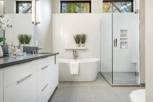 Modern bathroom with a white freestanding tub and a glass-enclosed shower. The space features white cabinets with dark countertops, light gray floor tiles, and a white textured wall behind the tub. Plants and decorative items add a fresh touch, illuminated by natural light from the newly completed window installation.