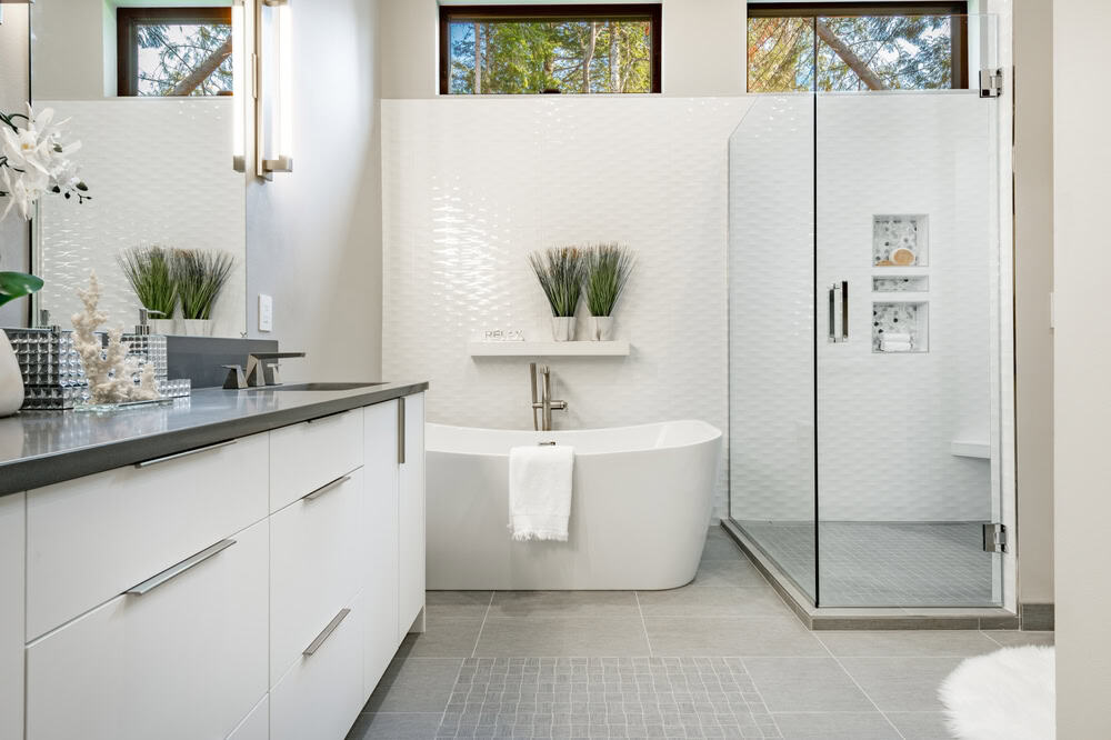 Modern bathroom with a white freestanding tub and a glass-enclosed shower. The space features white cabinets with dark countertops, light gray floor tiles, and a white textured wall behind the tub. Plants and decorative items add a fresh touch, illuminated by natural light from the newly completed window installation.