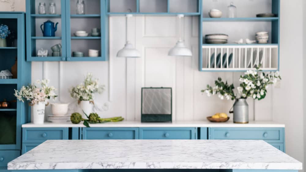 A bright kitchen features a white marble countertop island and blue cabinets with open shelves. The shelves display white dishes and various decorative items. There are two pendant lights above the counter, and flower arrangements adding a touch of nature, perfect for any homeowner considering a general contractor in Suffolk County.