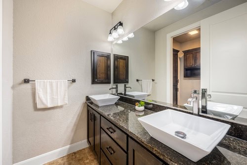 A modern bathroom, crafted by a General Contractor in Suffolk County, features a sleek black granite countertop with two white rectangular vessel sinks. Above the counter is a large mirror with three light fixtures. A hand towel hangs on the wall to the left, and a small decorative plant sits between the sinks.