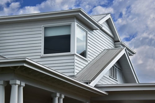 Close-up of a modern house with white siding and a gabled roof. The house boasts large, square windows on the second floor, ideal for those considering window installation. It features a covered porch with white columns. The sky is partly cloudy, providing a bright and airy background.
