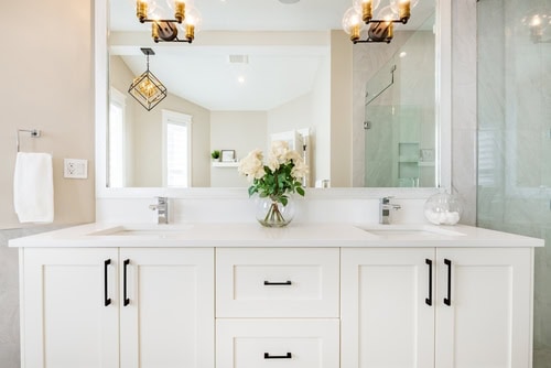 A bright and modern bathroom featuring a large mirror, dual white sinks with black hardware, and white cabinetry. A vase filled with white flowers is centered on the counter. Sconces with warm lighting flank the mirror, and a glass shower is visible on the right. Expert window installation adds natural light to complete the space.
