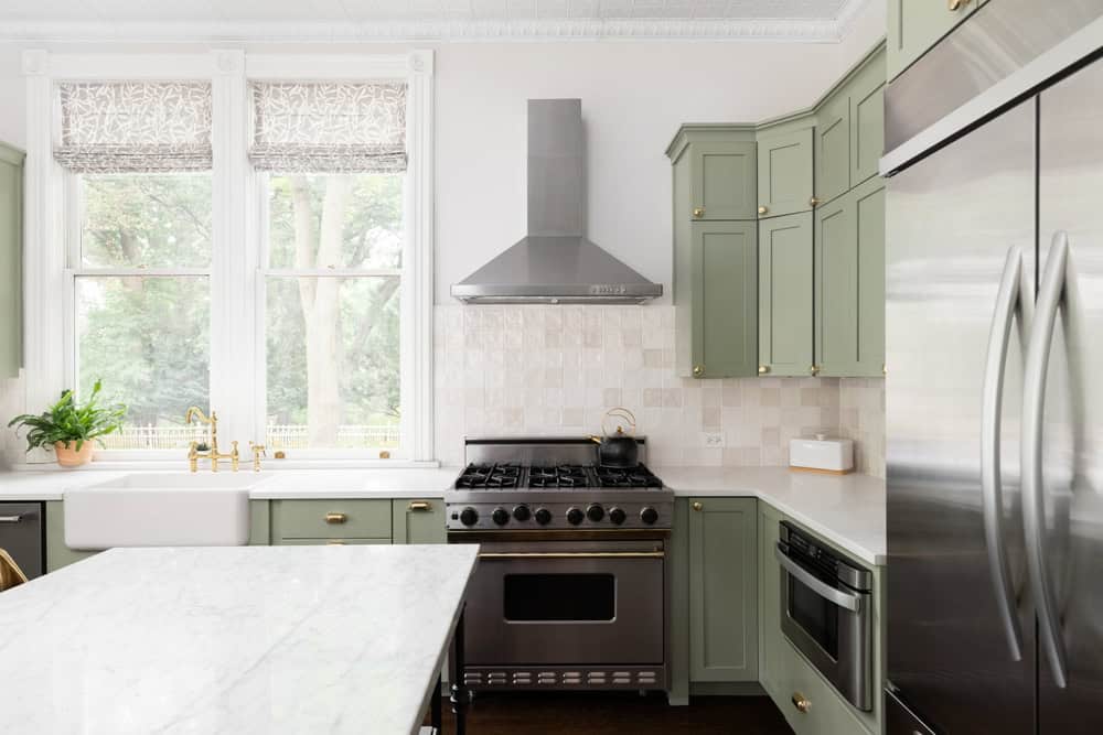 A modern kitchen featuring green cabinets with gold handles, a stainless steel stove with a range hood, and a farmhouse sink under two windows with patterned blinds installed during the Kitchen Remodeling in Suffolk County. A large refrigerator stands next to a marble island adorned with a potted plant.