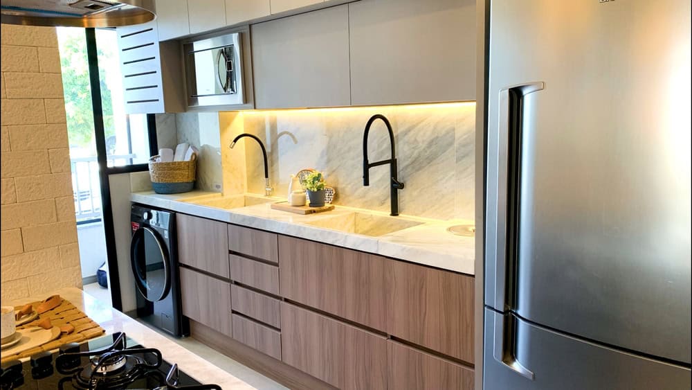 A modern kitchen features a marble countertop and backsplash with wooden lower cabinets and sleek grey upper cabinets. The kitchen is equipped with a stainless steel refrigerator, a black faucet, a built-in oven, and a washing machine. A window installation provides natural light.