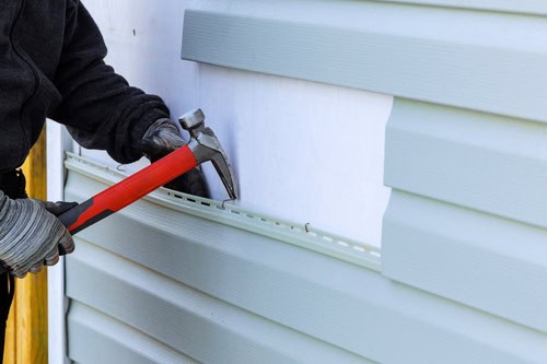 A person wearing work gloves uses a red and gray hammer to install or repair light green vinyl siding on a house, showcasing their expertise in window installation.