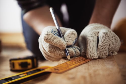 Close-up of a person wearing white knitted gloves, marking measurements on a wooden surface with a pencil and ruler. A yellow tape measure lies nearby. The scene suggests a detailed DIY home improvement project as a general contractor in Suffolk County.