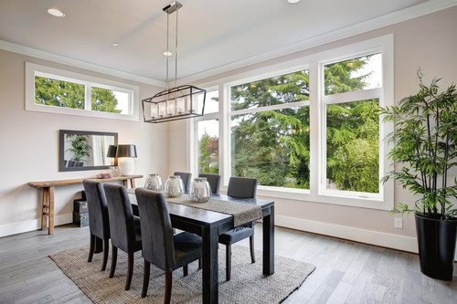 A modern dining room with a dark wooden table and six matching chairs. The table is set with glass decor on a gray runner. Large windows, recently upgraded by a general contractor through expert window installation, overlook lush greenery. A potted plant stands in the corner, and a console table with a lamp and mirror is against the wall.