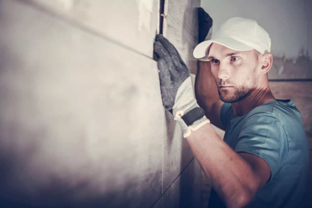 A man wearing a white cap and gloves carefully positions a tile on a wall, focused on ensuring it's aligned correctly. The wall appears to be partially tiled already, suggesting he might be working on a home renovation or kitchen remodeling project in Suffolk County.