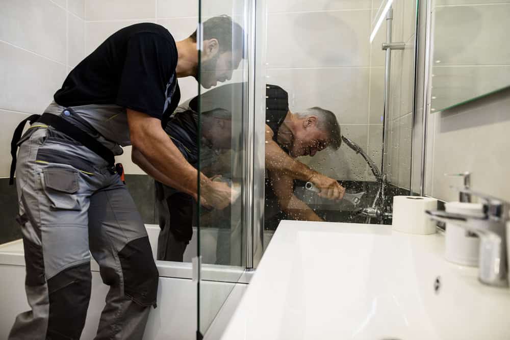 A plumber is working on a shower in a modern bathroom, fixing a pipe inside the shower enclosure. Another person, possibly an assistant, is holding a flashlight to provide additional light. The bathroom features a white sink, a mirror, and various tools—professionally handled by your trusted General Contractor Suffolk County experts.