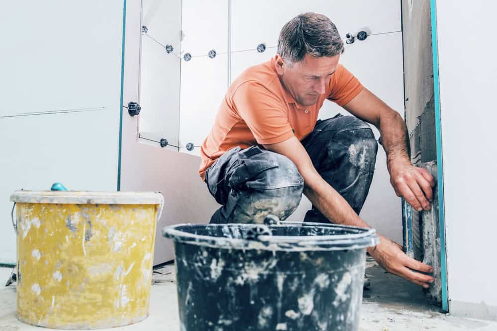 A man in a construction outfit, wearing a bright orange polo shirt and dirty work pants, is tiling a wall. He is placing a tile onto the adhesive. In the foreground, there are two buckets, one yellow and one black, used for mixing materials— showcasing the expertise of a Bathroom Remodeler in Suffolk County.