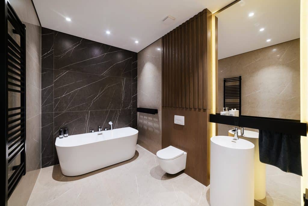 A modern bathroom in Suffolk County with a freestanding white bathtub, a wall-mounted toilet, and a cylindrical pedestal sink. The walls feature dark marble tiles and light beige tiles. The room has recessed lighting and a sleek black towel radiator next to the bathtub—all crafted by a top General Contractor Suffolk County.