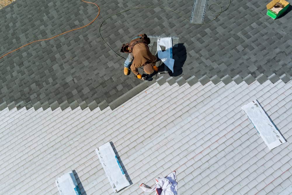 A person is installing shingles on a roof. The top part of the roof has green shingles, while the lower part is covered with white tiles. Nearby, rolls of roofing material and tools are scattered on the white tiles. An orange extension cord is visible, perfect for your next Roof Repair Suffolk County project.