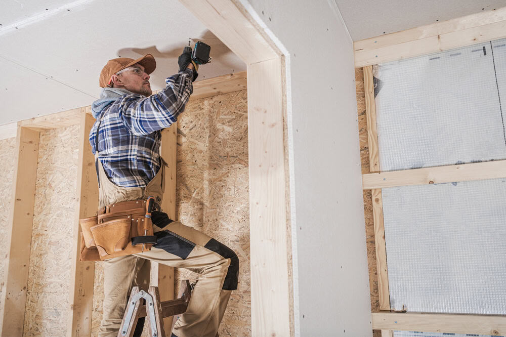 A construction worker wearing a plaid shirt, hat, and tool belt is standing on a ladder while using a drill to work on the ceiling of a partially constructed room with exposed wooden frames and insulation, likely part of a kitchen remodeling project in Suffolk County.