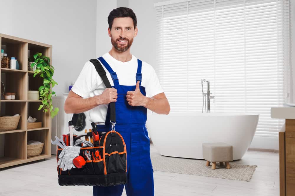 A smiling man wearing blue overalls and a white t-shirt stands in a modern bathroom, giving a thumbs-up. He holds a tool bag filled with various tools. The bathroom remodeler from Suffolk County admires the space, which features a freestanding bathtub, a plant, and shelves with towels and baskets.