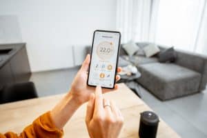 A person is using a smartphone to control a home thermostat. The screen displays a temperature setting of 22.0°C for the living room, along with options for controlling different zones. In the background, a modern living room with a gray sofa, kitchen, and recently completed window installation is visible.