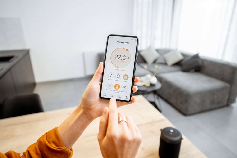 A person is using a smartphone to control a home thermostat. The screen displays a temperature setting of 22.0°C for the living room, along with options for controlling different zones. In the background, a modern living room with a gray sofa, kitchen, and recently completed window installation is visible.