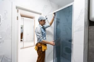 A man wearing a cap and a tool belt is installing a glass shower door in a modern bathroom with white marble walls. The bathroom has a clean, minimalistic design, and an open door with a mirror is visible in the background. This skilled general contractor in Suffolk County ensures precision craftsmanship.