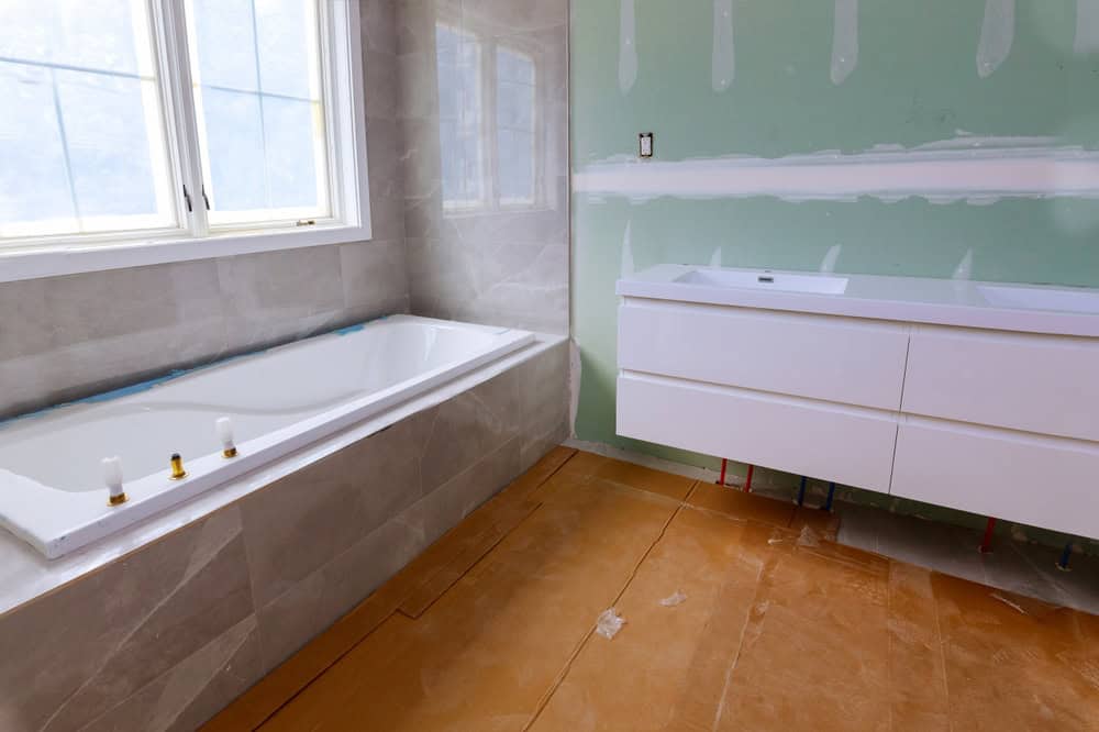 A bathroom mid-renovation by General Contractor Suffolk County features a new white bathtub along the left wall, with unpainted drywall and patches visible. On the right, a white double-sink vanity is installed. The floor is covered with protective paper over tiles.