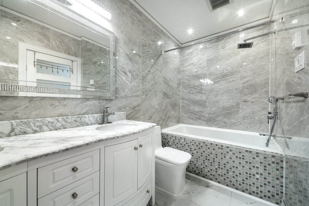 A luxurious bathroom designed by a top Bathroom Remodeler in Suffolk County features marble walls and flooring, a white vanity with a marble countertop and sink, a large mirror with lights above it, a white toilet, and a bathtub with a mosaic tile accent. Recessed lighting and a sleek showerhead complete the modern design.