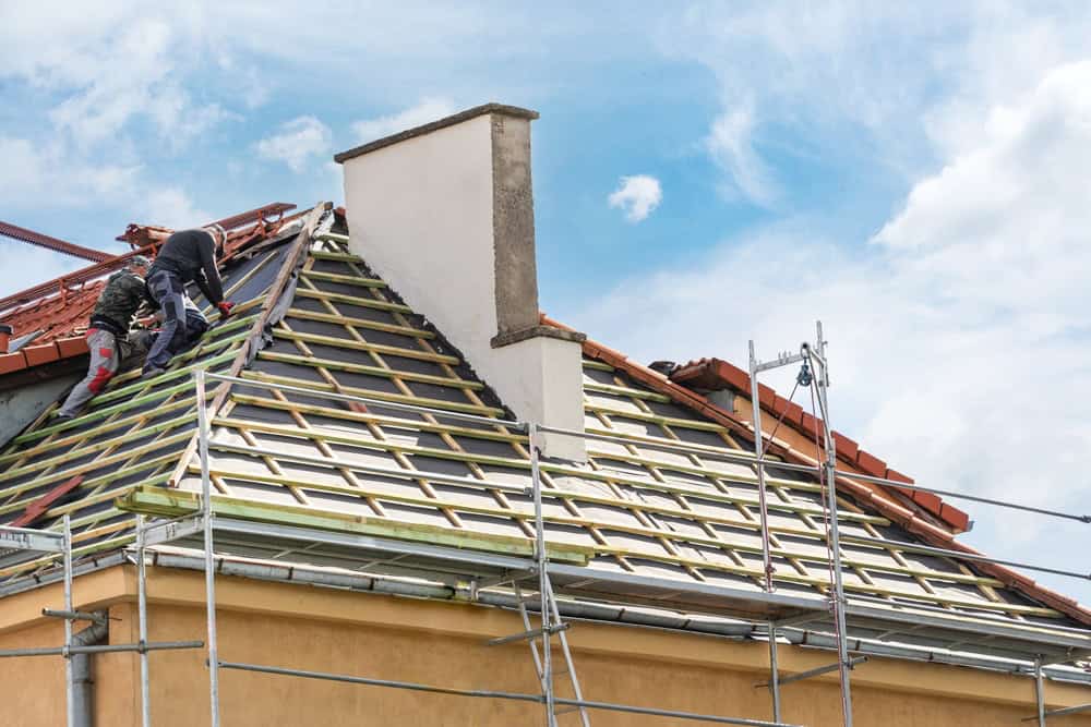 Two workers climb on a roof with visible framework, replacing tiles. Supported by scaffolding around the house, they work diligently under a partly cloudy sky. Their safety harnesses ensure a secure working environment—a routine task for any experienced general contractor in Suffolk County.