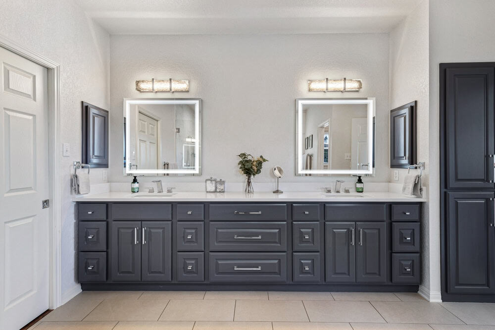 A modern bathroom with two lighted mirrors above a double sink vanity featuring dark gray cabinets, designed by a top Bathroom Remodeler in Suffolk County. The white countertops have soap dispensers and other small decor items. Bright light fixtures are mounted above each mirror, and there's a door on the left.