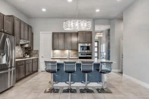 A modern kitchen features stainless steel appliances, grey cabinets, a center island with a light grey countertop, and four transparent barstools. An elegant circular light fixture hangs above the island. The floor is tiled, and the walls are painted a soft grey. Nearby windows showcase expert window installation.