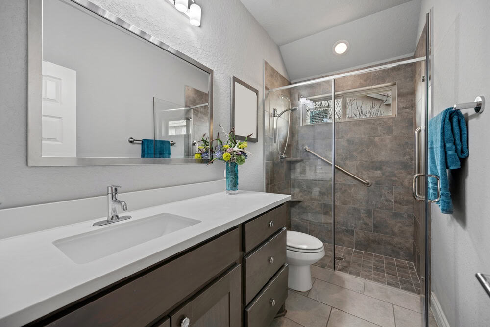 A modern bathroom, designed by a General Contractor in Suffolk County, features a single-sink vanity with a large mirror, floral decorations, and a glass-enclosed shower. The shower boasts gray tiles and a built-in seat, while blue towels hang on racks. The overall decor is sleek and neutral-toned.