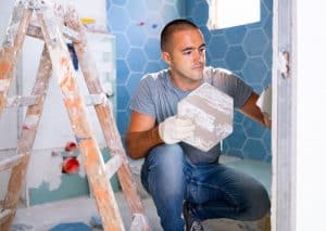 A man in a grey t-shirt and blue jeans is kneeling in a partially tiled bathroom, holding a piece of hexagonal tile as he installs it on the wall. Beside him, a wooden ladder with paint marks stands ready for further tasks like siding Suffolk County or window installation. Bathroom fixtures are visible around him.