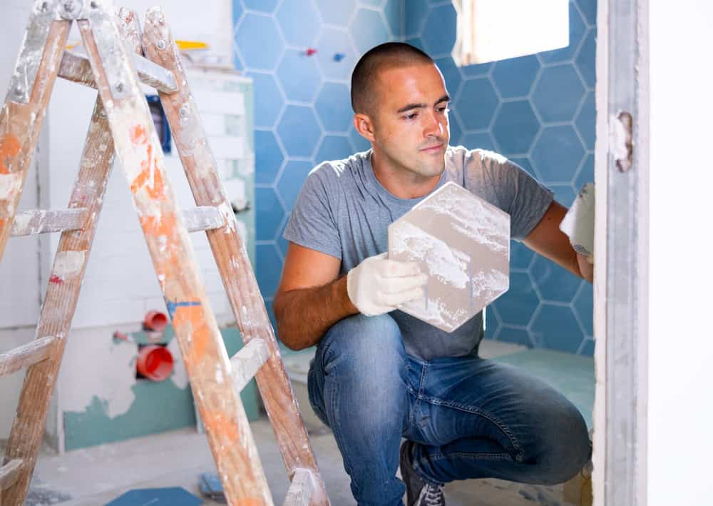 A man in a grey t-shirt and blue jeans is kneeling in a partially tiled bathroom, holding a piece of hexagonal tile as he installs it on the wall. Beside him, a wooden ladder with paint marks stands ready for further tasks like siding Suffolk County or window installation. Bathroom fixtures are visible around him.