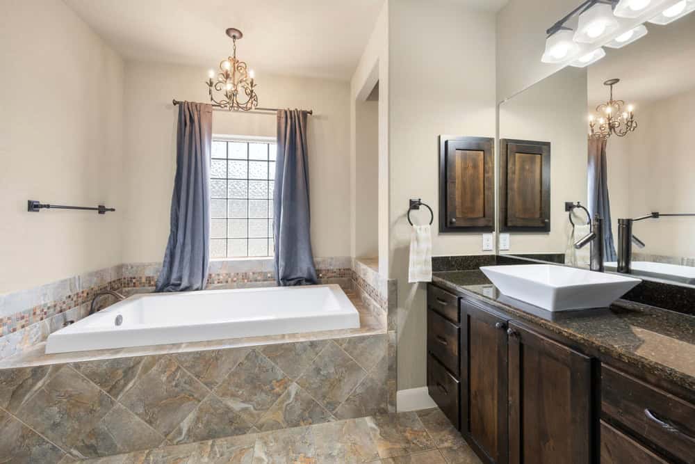 A modern bathroom features a large bathtub with tiled surroundings under a newly installed window with grey curtains. There are dark wooden cabinets with a granite countertop, a white vessel sink, and a large mirror with light fixtures mounted above. A chandelier hangs from the ceiling.