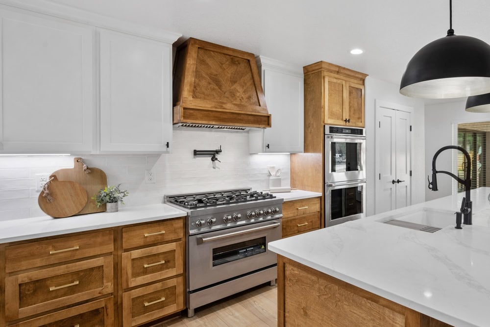 A modern kitchen featuring wooden cabinets, white countertops, and stainless steel appliances, including a large stove with a wooden range hood and a built-in oven. A kitchen island with a black faucet and two pendant lights overhead is also present, showcasing the craftsmanship of General Contractor Suffolk County.