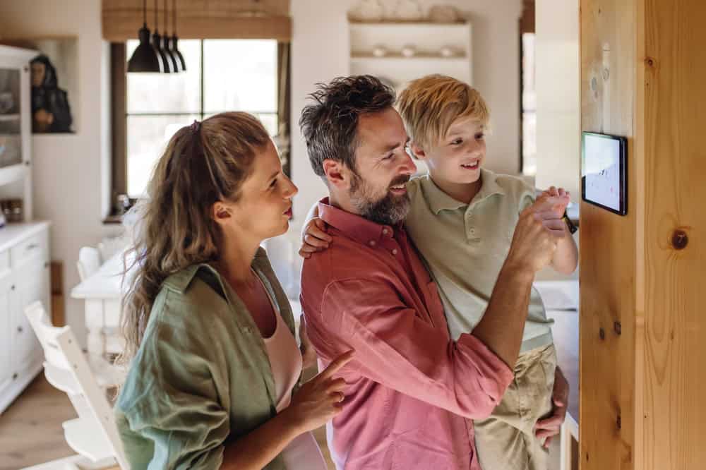 A woman, man, and young boy stand together around a smart home device mounted on a wall. The man, in a red shirt, and the boy, dressed casually, touch the device while the woman, in a light green shirt, looks on. Through the window installation behind them, they appear cheerful in a bright, modern kitchen.