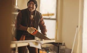 A person wearing safety glasses, a beanie, gloves, and a plaid shirt is smiling while using a hand saw to cut a piece of wood in a workshop. The background includes tools and equipment used for tasks like kitchen remodeling in Suffolk County, with light filtering in through the windows.