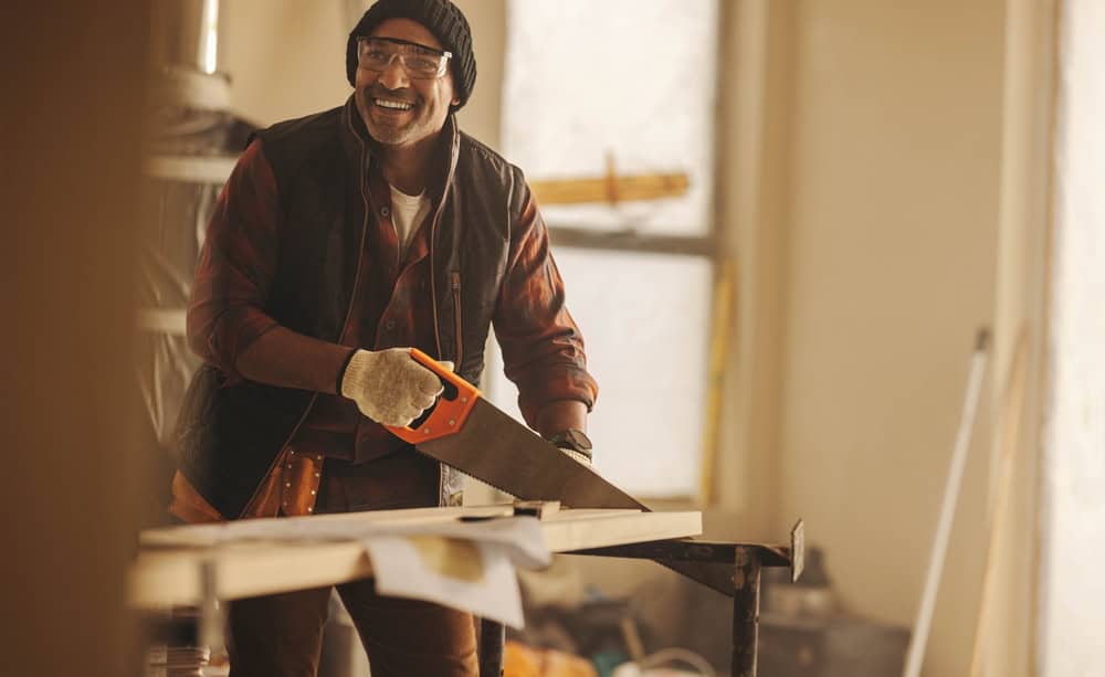 A person wearing safety glasses, a beanie, gloves, and a plaid shirt is smiling while using a hand saw to cut a piece of wood in a workshop. The background includes tools and equipment used for tasks like kitchen remodeling in Suffolk County, with light filtering in through the windows.