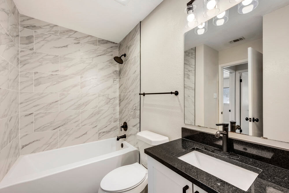 A modern bathroom by a Bathroom Remodeler in Suffolk County featuring a white bathtub with marble-patterned tiles, a toilet, and a black countertop sink with white cabinets. The sink area includes a large mirror and wall lighting fixtures. The room is well-lit and has a neutral color scheme.