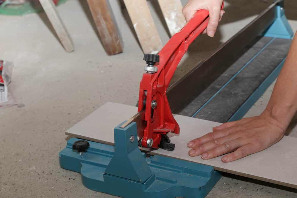 A General Contractor in Suffolk County uses a manual tile cutter to cut a piece of tile. The red cutting handle is pressed down on the tile, positioned on the blue base of the cutter, while the other hand stabilizes it.