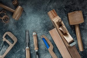 A set of woodworking tools including a wooden mallet, a hand plane, chisels, a coping saw, and wood shavings arranged on a textured gray surface. The tools have wooden handles or parts, showing a polished and well-crafted finish—perfect for any kitchen remodeling project in Suffolk County.