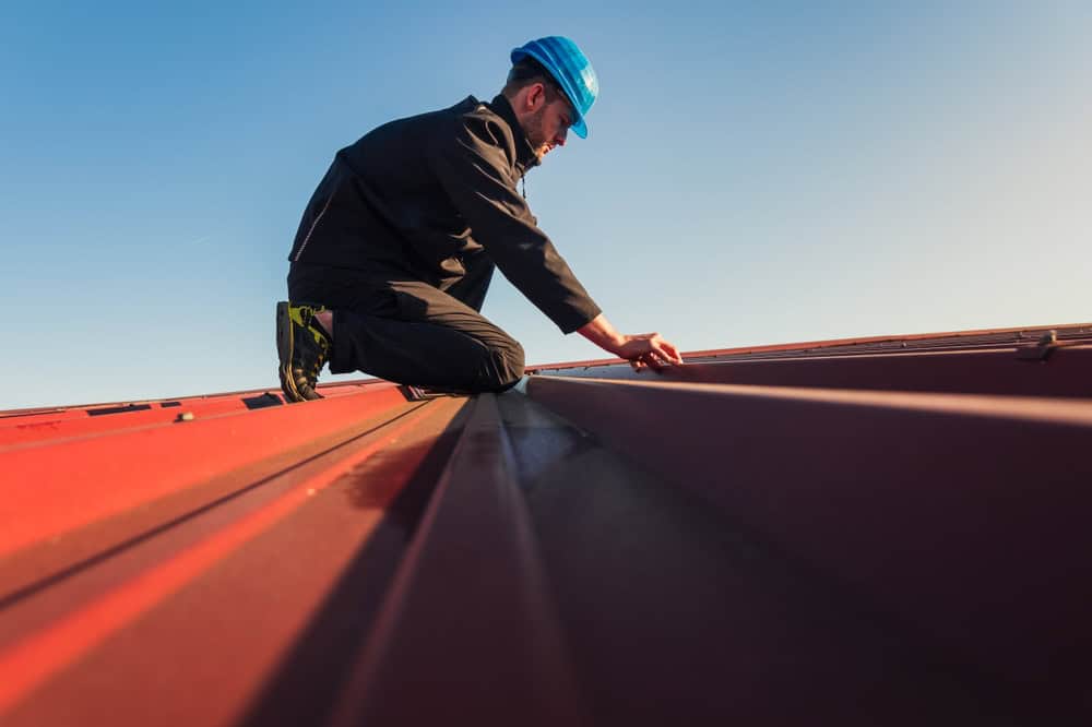 A person in work clothes and a blue hard hat is kneeling on a red metal roof, inspecting or performing maintenance. The sky is clear and bright in the background, showcasing the dedication of this General Contractor Suffolk County professional.