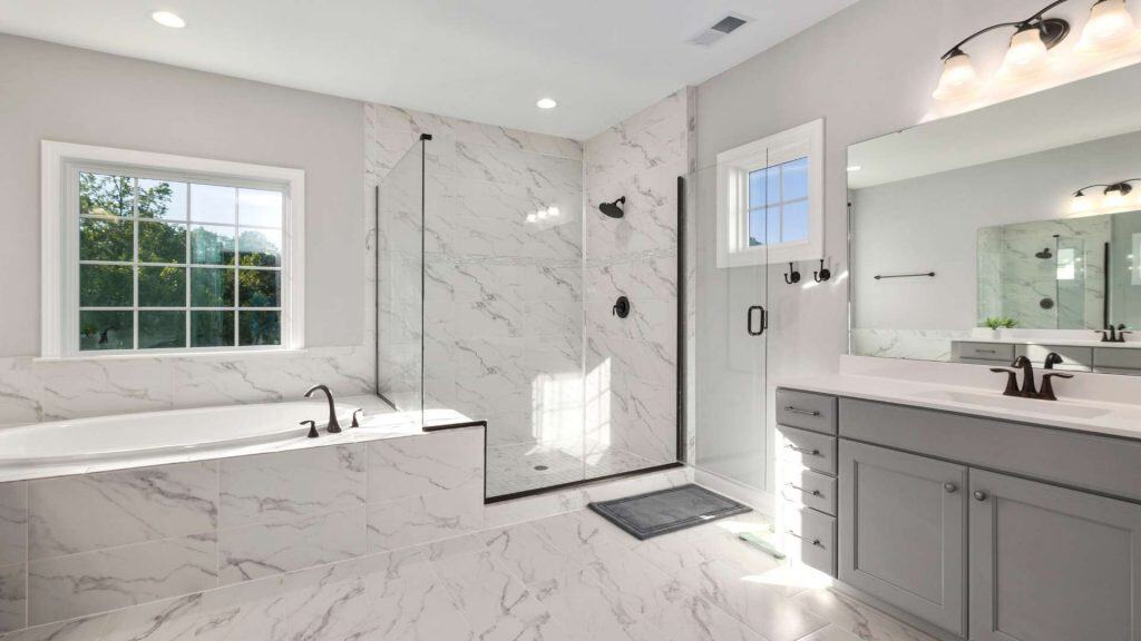 A modern bathroom with a marble-tiled floor and walls, featuring a glass-enclosed shower, a large bathtub by a window, a gray vanity with double sinks, and black fixtures. A small mat lies in front of the shower.
