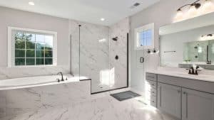 A modern bathroom with a marble-tiled floor and walls, featuring a glass-enclosed shower, a large bathtub by a window, a gray vanity with double sinks, and black fixtures. A small mat lies in front of the shower.