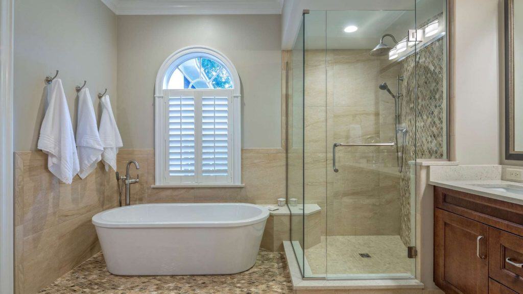 A bathroom featuring a freestanding white bathtub with three towels hanging on hooks next to it. There's a glass-enclosed shower with a tiled accent wall and a window with shutters above the tub. A wooden vanity is partially visible.