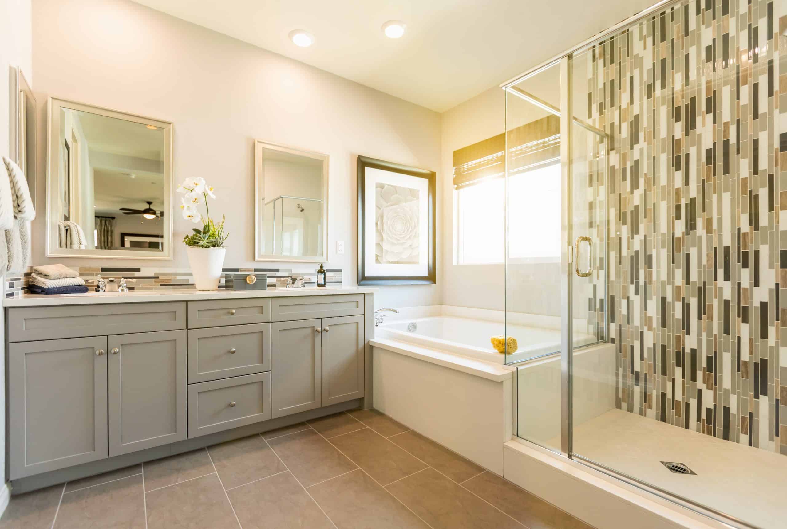 Modern bathroom with gray cabinetry, two mirrors, and a large bathtub. The shower features a glass door and mosaic tile design. Natural light streams in through a window with blinds. Perfect for those considering bathroom remodeling, the space is enhanced by a potted orchid and framed artwork.
