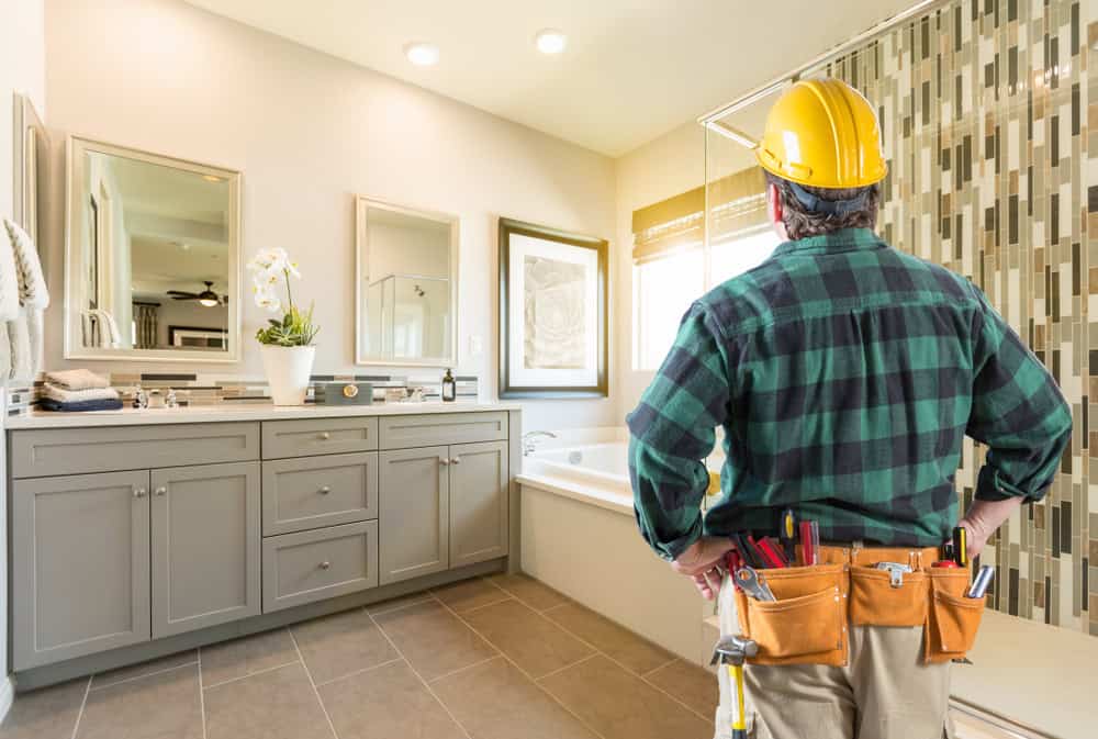 A construction worker wearing a yellow hard hat and tool belt stands in a modern bathroom. The room has a double sink with mirrors, a bathtub, and a decorative tiled wall. Natural light streams in through a window with blinds.